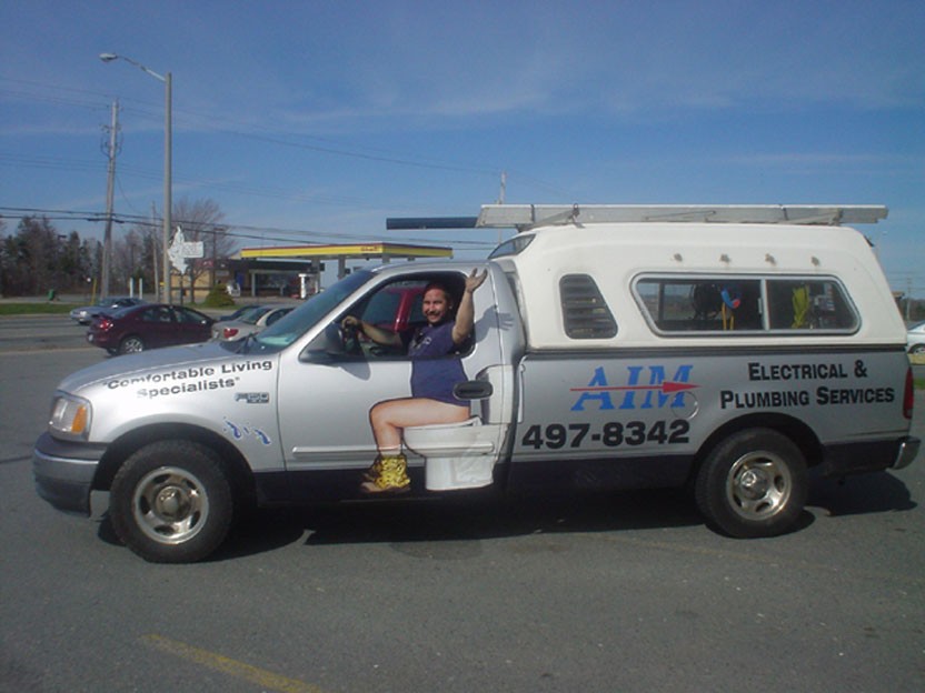 Silver pickup truck with a toilet in the driver's seat and plumbing company decals