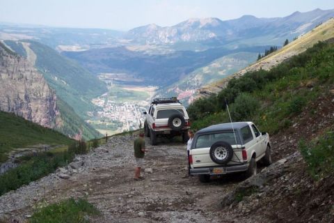 1990 Ford Bronco - Two Ford Broncos navigate a challenging mountain trail with a scenic valley below.
