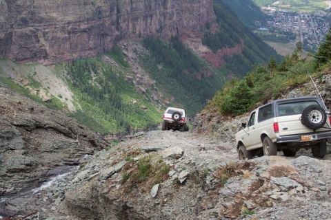 1990 Ford Bronco - Two Ford Broncos navigate a steep, rocky mountain pass with a town visible in the distance.