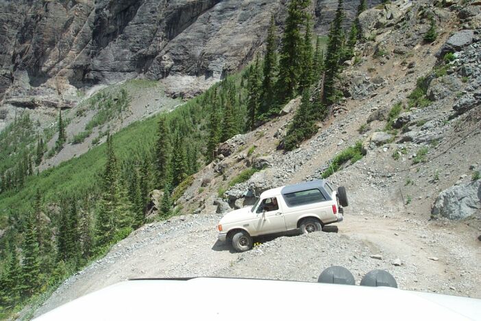 White SUV on a narrow, rocky mountain road with steep, forested slopes