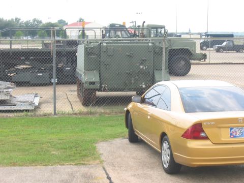 1988 Ford Bronco - A gold Honda Civic coupe is parked in front of a military truck behind a chain-link fence.