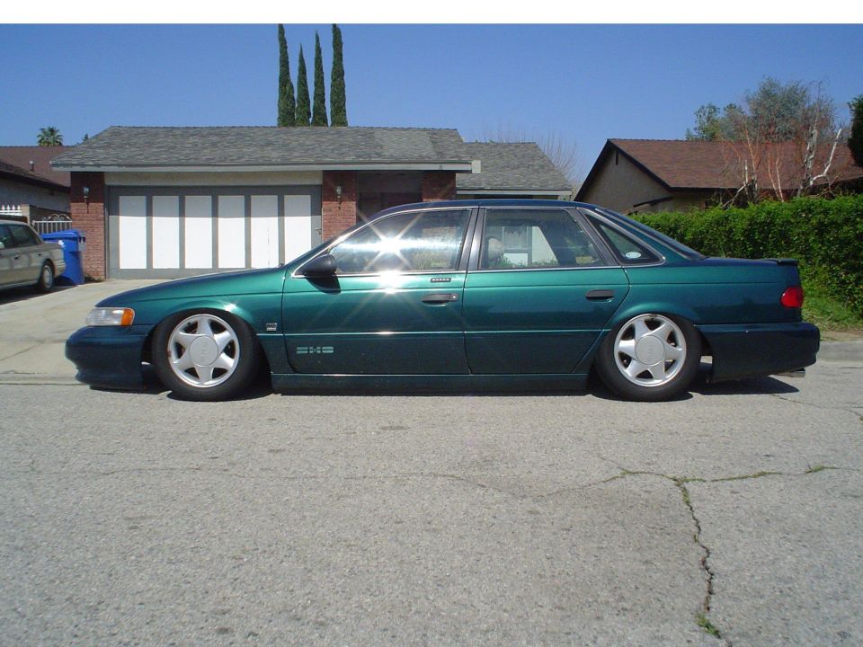 Heavily lowered green Ford Taurus SHO sedan in profile at a curb with cypress trees behind