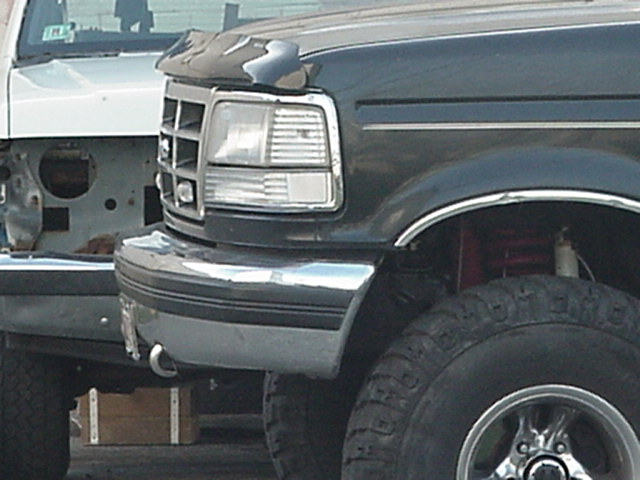 Dark truck front corner with chrome grille and bumper. Another white truck is visible behind