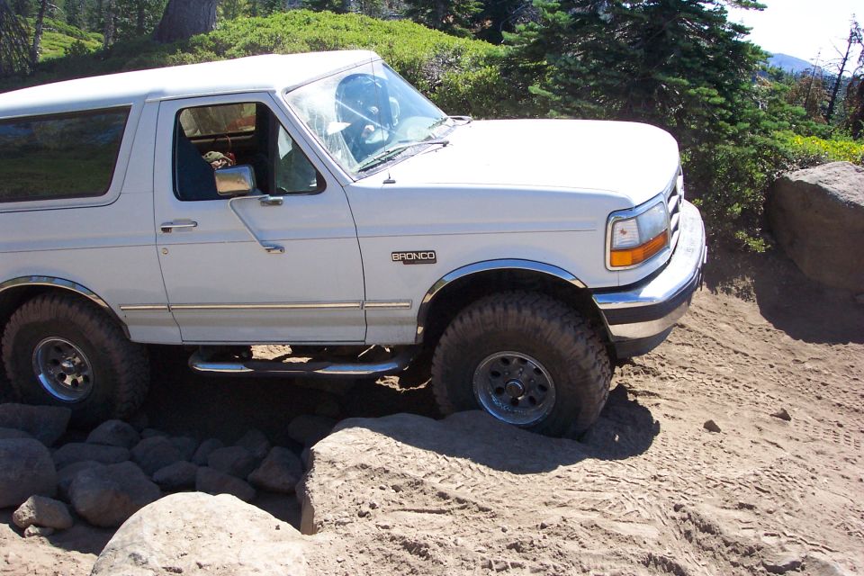 White Ford Bronco with lift and off-road tires crawling over rocks on a dirt trail, passenger side view