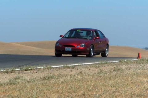 1997 Ford Taurus - A red 1997 Ford Taurus navigates a turn on a racetrack, indicated by the number 65 on its windshield.