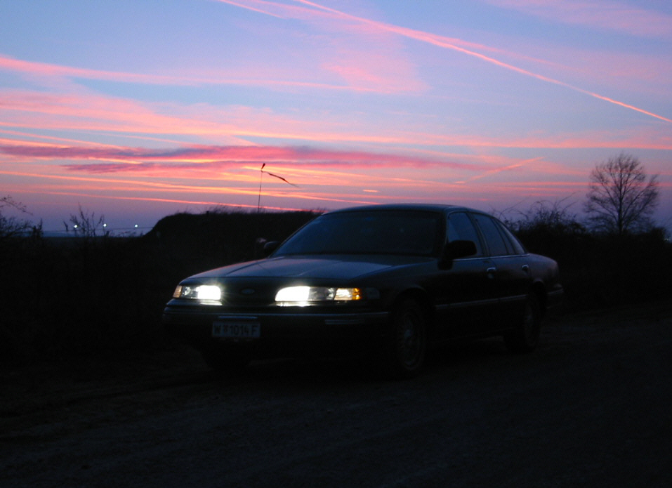 1992 Ford Crown Victoria parked on dirt road with headlights on, sunset sky