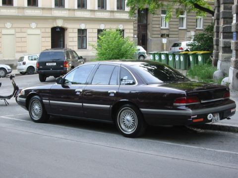 1992 Ford Crown Victoria - A dark 1992 Ford Crown Victoria sedan is parked on a street, with other vehicles and buildings in the background.
