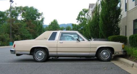 1985 Ford LTD Crown Victoria - A tan/beige two-door Ford LTD Crown Victoria coupe photographed in profile, showing its wire-spoke wheel covers, chrome trim, and vinyl-loo…