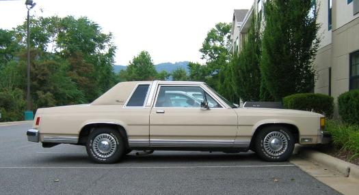 Beige two-door Ford LTD Crown Victoria coupe seen in side profile in a parking lot.