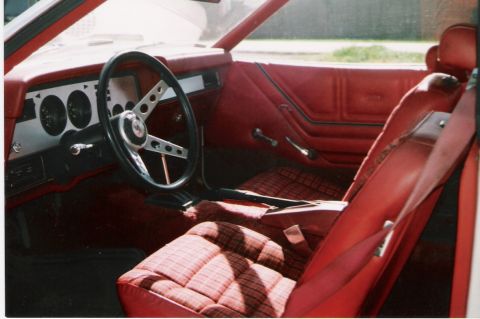 1978 Ford Mustang II - The interior of a vintage car features red vinyl seats with plaid inserts and a dashboard with round gauges.