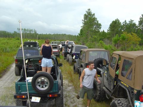 1993 Ford Bronco - A group of off-road vehicles are parked on a dirt road surrounded by trees and brush.