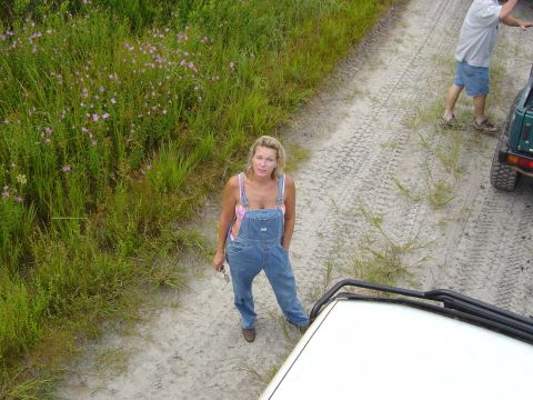 1993 Ford Bronco - A woman in denim overalls stands on a sandy dirt road, with two off-road vehicles nearby and a Supermotors.net logo in the foreground.