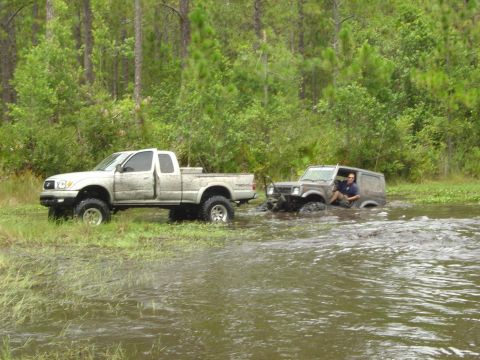 1993 Ford Bronco - A gray SUV is stuck in the water while a white pickup truck attempts to tow it out.