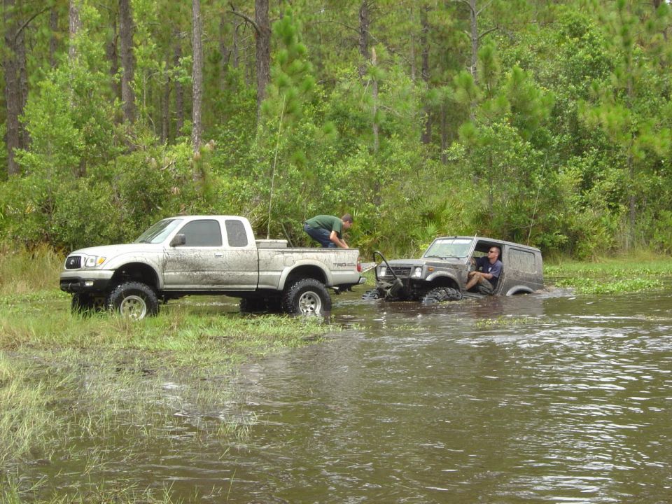 White pickup truck towing a stuck SUV in muddy water, surrounded by trees