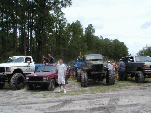 1993 Ford Bronco - A group of people stand around several heavily modified off-road vehicles in a wooded area.