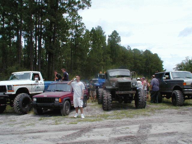 Several lifted off-road vehicles parked in a dirt lot with trees in the background