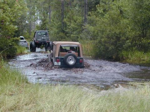 1993 Ford Bronco - Multiple Jeeps are seen driving through a muddy creek in a forest environment.