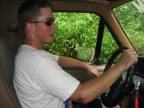 1988 Ford Bronco - A man wearing sunglasses drives a vehicle, with dense green foliage visible through the window.