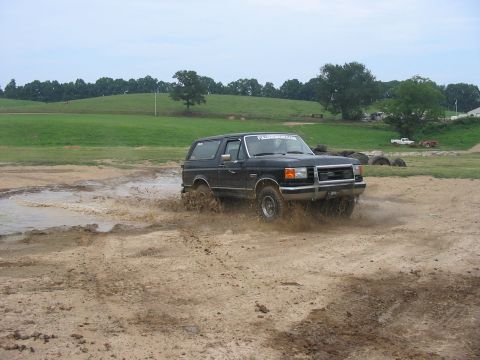 1988 Ford Bronco - A black 1988 Ford Bronco navigates a muddy track, kicking up water and dirt.