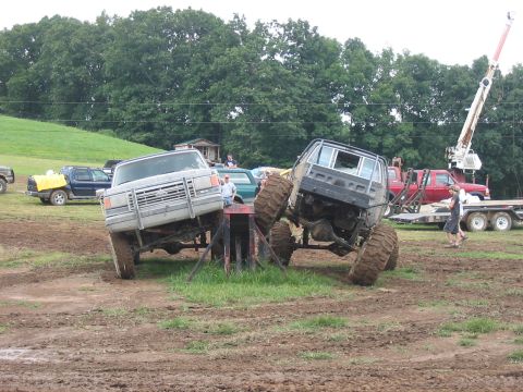 1988 Ford Bronco - Two heavily modified trucks are positioned in a muddy outdoor area with trees in the background.