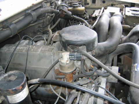1992 Ford Bronco - Close-up of a dusty engine bay showing a black round air cleaner housing, corroded metal components, and multiple black rubber vacuum hoses…