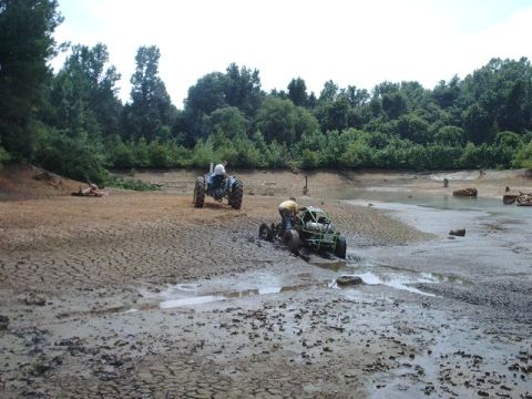 1993 Ford Bronco - Two off-road vehicles are stuck in the mud at the bottom of a dry lake bed, with trees in the background.
