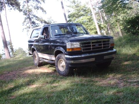 1993 Ford Bronco - A dark-colored Ford Bronco is parked on a grassy incline surrounded by trees.