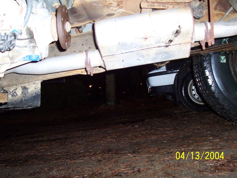 Underside view of a vehicle's exhaust pipe and muffler, with another car visible underneath