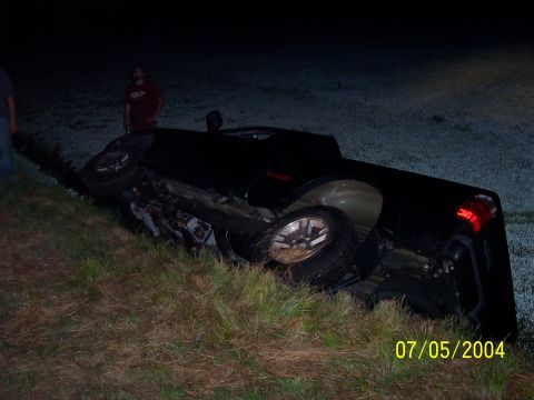 1994 Ford Bronco - A black pickup truck lies overturned on its side in a grassy area at night, with two individuals observing the scene.
