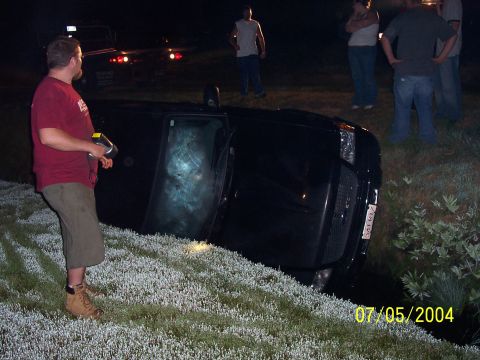 1994 Ford Bronco - A black SUV has flipped onto its side in a grassy ditch, with onlookers gathered around.