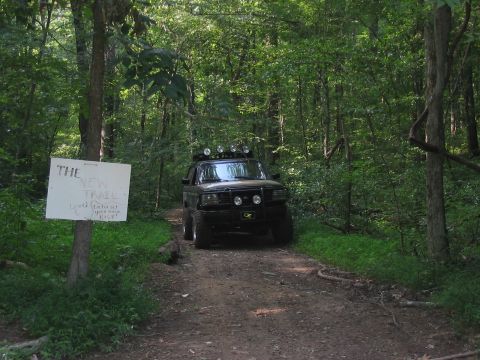 1988 Ford Bronco - A black 1988 Ford Bronco navigates a dirt path through a dense forest, marked by a sign indicating 'The New Trail'.