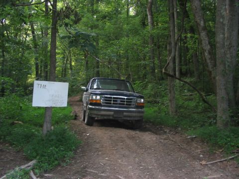 1988 Ford Bronco - SUV drives down a wooded dirt road next to a sign indicating a new trail.