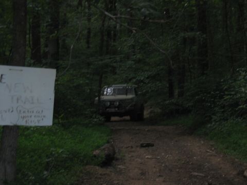 1988 Ford Bronco - A tan off-road vehicle drives down a wooded trail past a sign warning of risk.