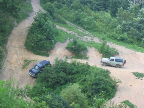 1988 Ford Bronco - Two SUVs navigate a winding dirt path through a lush, green, wooded landscape.
