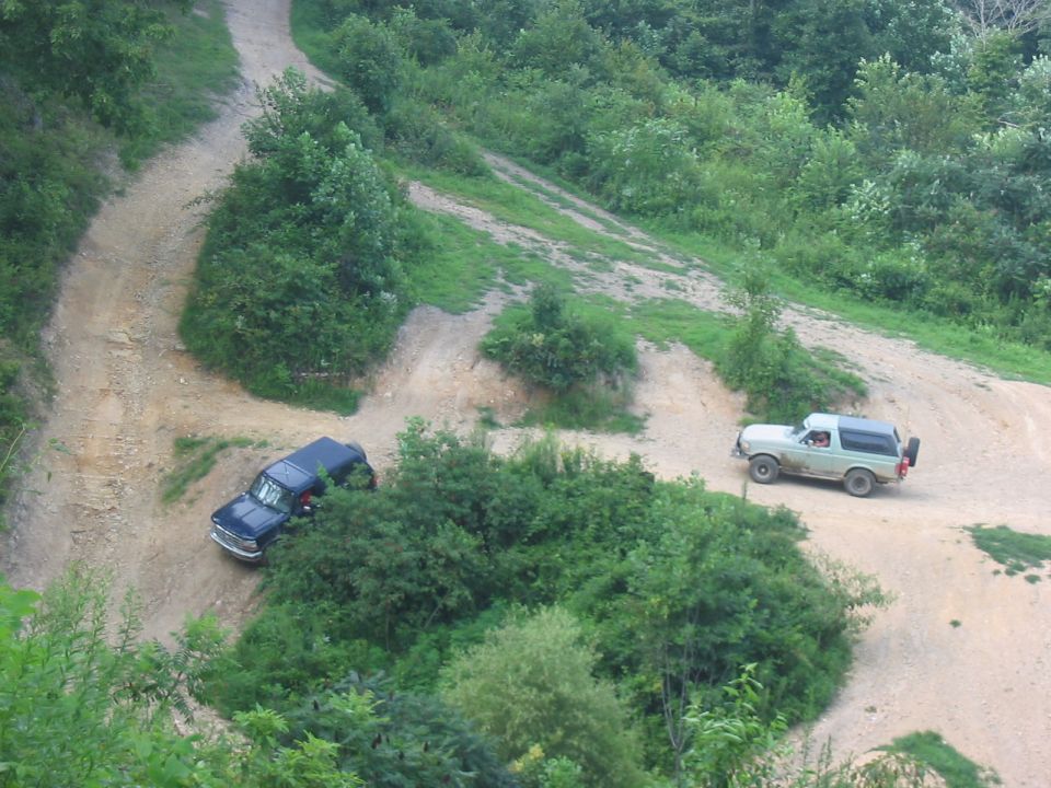 Two SUVs driving on a dirt road in a wooded area. One is a light-colored Ford Bronco, the other is a dark SUV