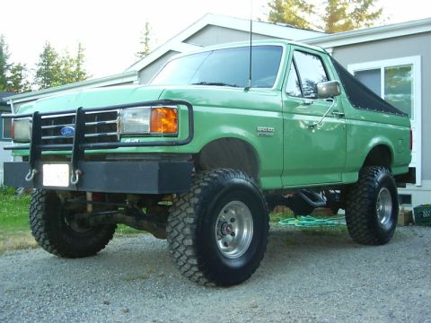 1990 Ford Bronco - Three-quarter front driver-side view of a lifted mint-green Ford Bronco parked on gravel next to a house, with the black soft top folded do…