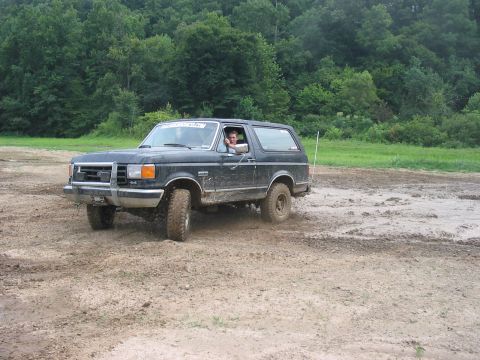 1988 Ford Bronco - A dark-colored Ford Bronco navigates a muddy terrain with trees lining the horizon.