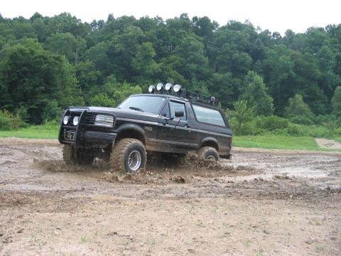 1988 Ford Bronco - This black Ford Bronco is navigating a challenging muddy terrain, kicking up water and dirt.