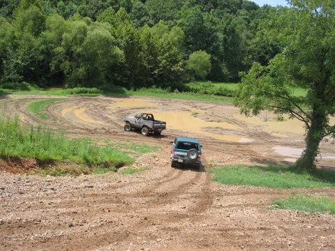 1988 Ford Bronco - Two trucks navigate a challenging muddy track surrounded by lush green trees.