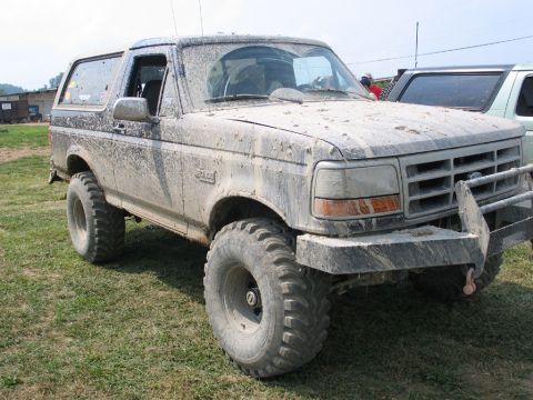 1995 Ford Bronco - A heavily muddied 1992 Ford Bronco sits on grassy terrain, its off-road tires prominent.
