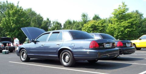 1998 Ford Crown Victoria - Several cars are parked in a lot, some with their hoods open, under a bright sky with trees in the background.