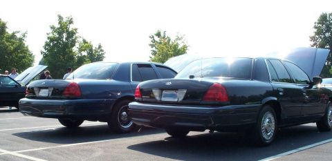 1998 Ford Crown Victoria - Two Ford Crown Victorias are seen from the rear in a parking lot, with one vehicle's hood open.