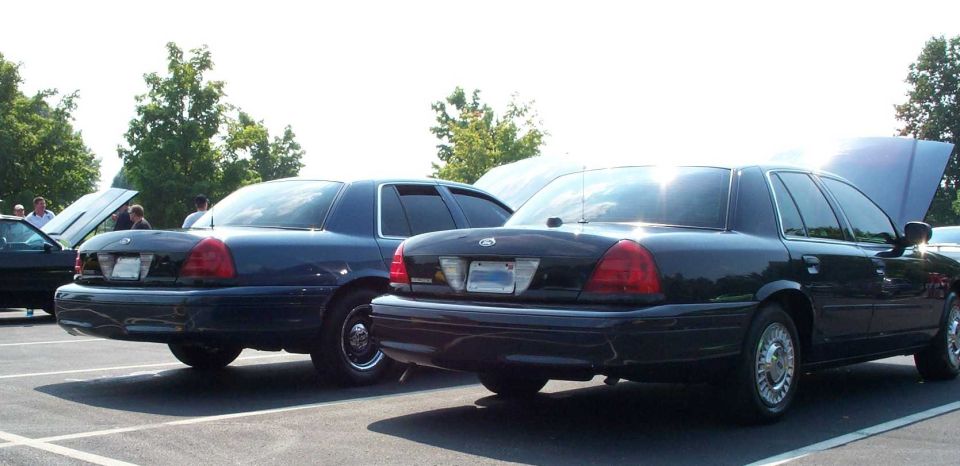Rear view of two dark blue Ford Crown Victoria sedans parked side by side at a car meet