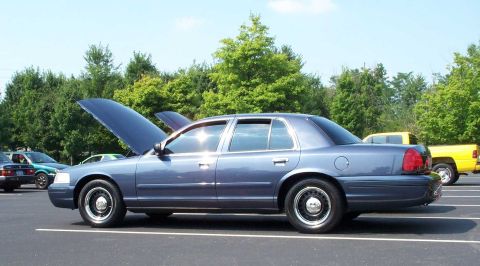 1998 Ford Crown Victoria - A blue 1998 Ford Crown Victoria is parked with its hood open at an outdoor car meet.