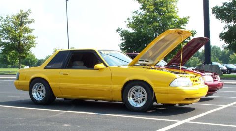 1998 Ford Crown Victoria - A yellow coupe with its hood open and aftermarket wheels is parked at an outdoor event.