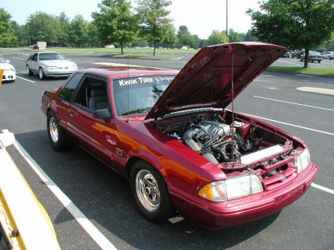 1998 Ford Crown Victoria - A red Ford Mustang is parked with its hood up, revealing the engine. Another car is visible in the background.