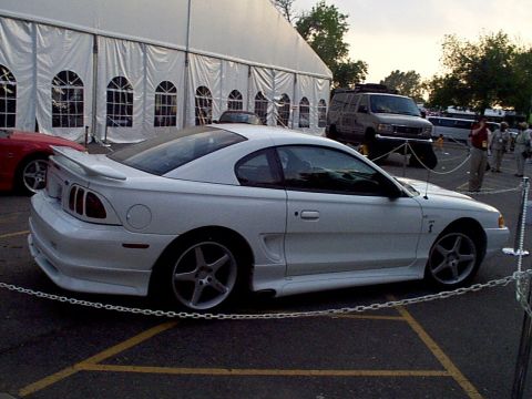 1999 Ford Taurus - Side view of a white Ford Mustang coupe, parked outdoors near a large tent.