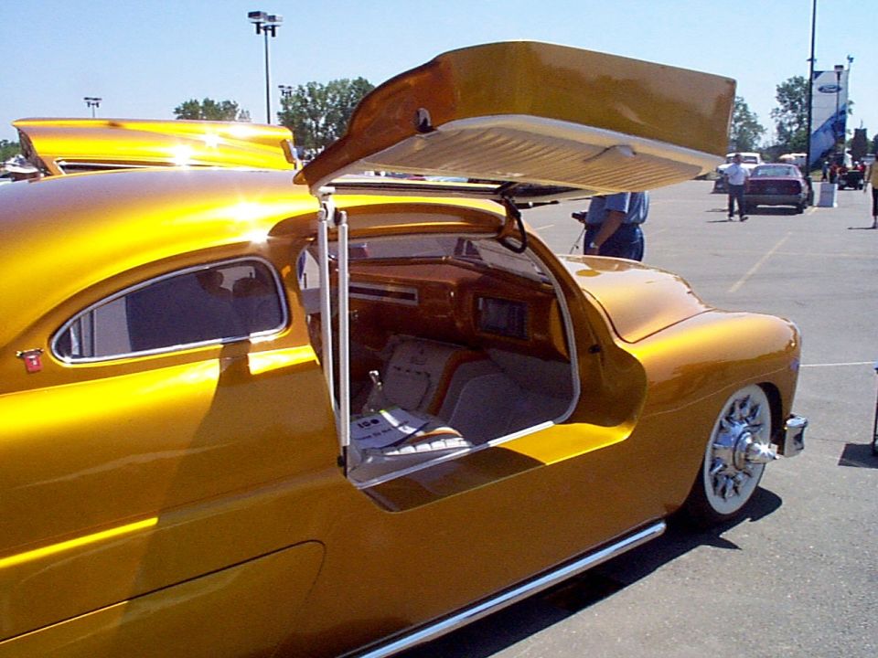 Customized classic car with gold paint, gullwing doors open, showing wood-paneled interior and white-wall tires