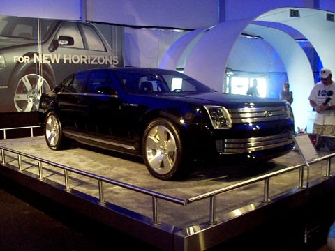1999 Ford Taurus - A dark blue concept car is presented on a display platform with a backdrop reading 'FOR NEW HORIZONS'.