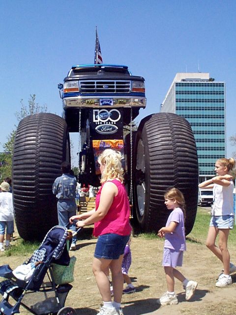 1999 Ford Taurus - A massive Ford monster truck is displayed with an American flag on top, drawing attention from onlookers.